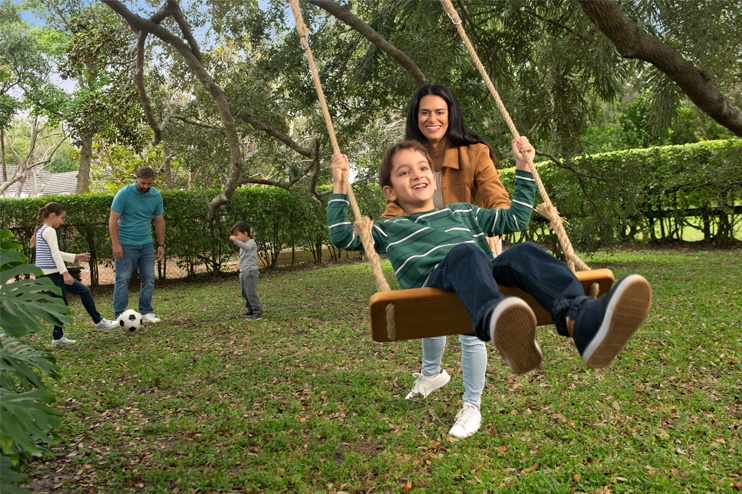 A mother pushes her son on a swing while her husband plays soccer on the lawn with her two other children.