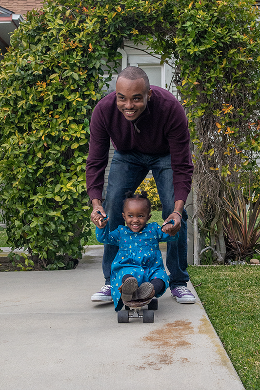 Father holding toddler girl's hands as she sits on a skateboard outside