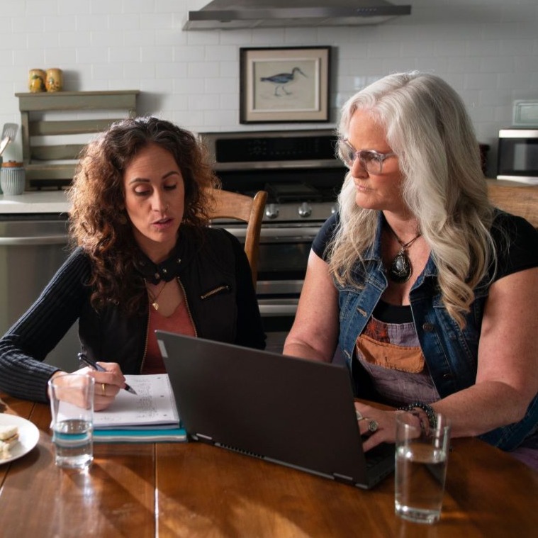 Two women sit at a kitchen table and look at a laptop to review a financial offering from their credit union.