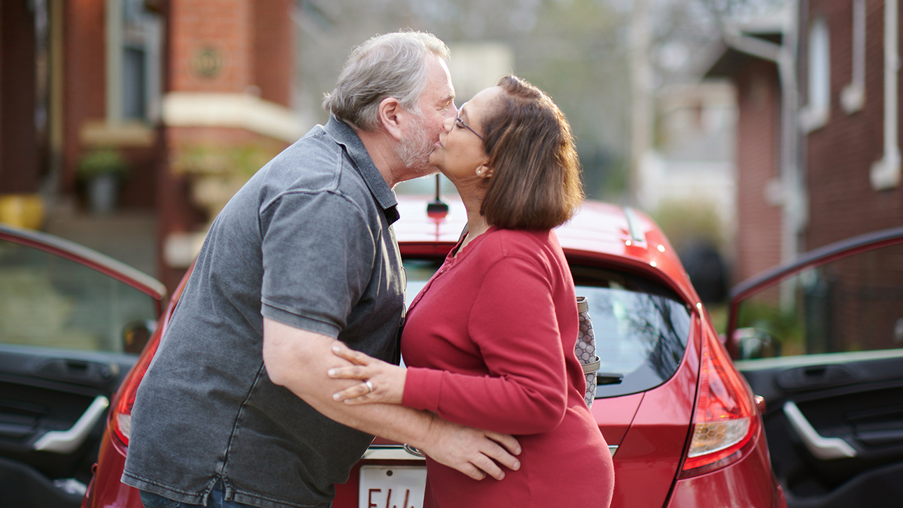 Man and woman embracing, about to kiss other’s cheek, in front of a car with open doors