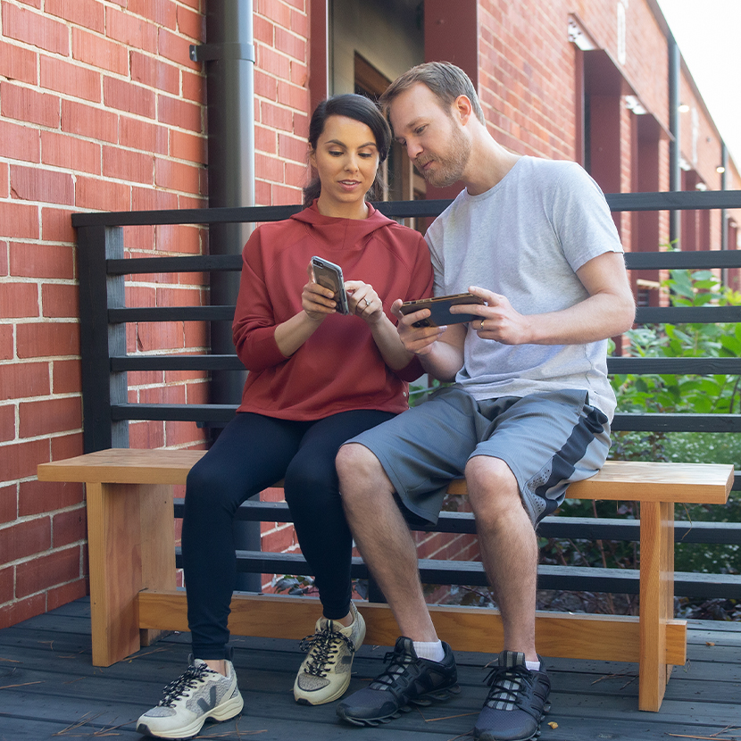 A man and a woman sitting together on a bench outside looking at their smartphones