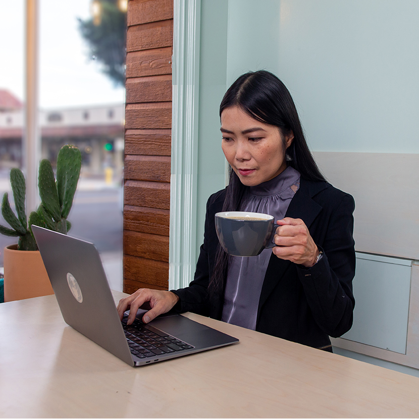 Woman in a coffee shop sitting at a table looking at her laptop and holding a coffee.