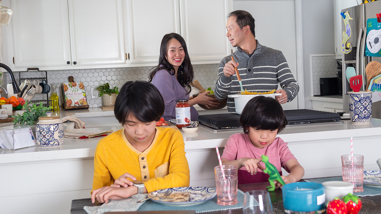 Parents cooking together in the kitchen while their kids sit at the dining table