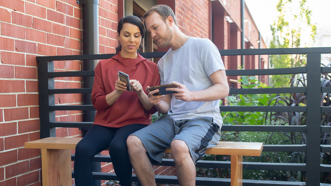 A man and a woman sitting together on a bench outside looking at their smartphones