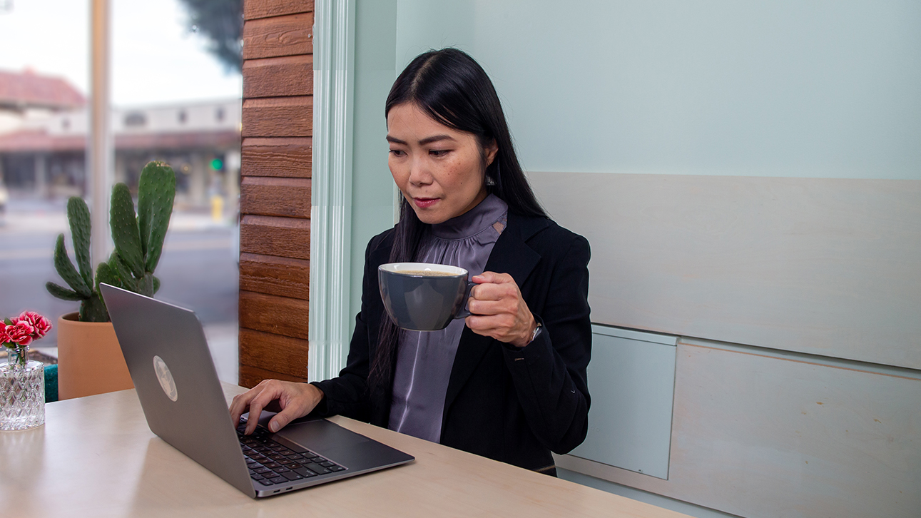 Woman in a coffee shop sitting at a table looking at her laptop and holding a coffee.