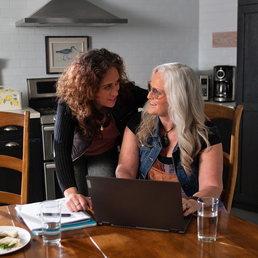 Two women sit at a kitchen table and look at a laptop to review a financial offering from their credit union.