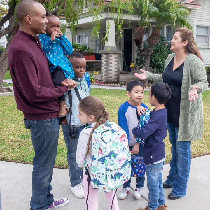 Parents stand on a sidewalk with children in front of a house as they wait for a school bus.