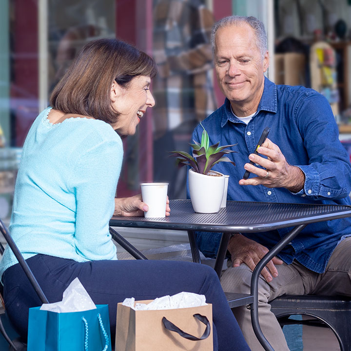 A husband and wife seated in an outdoor cafe use a cell phone to make a TruStage Policy Payment from their online account.