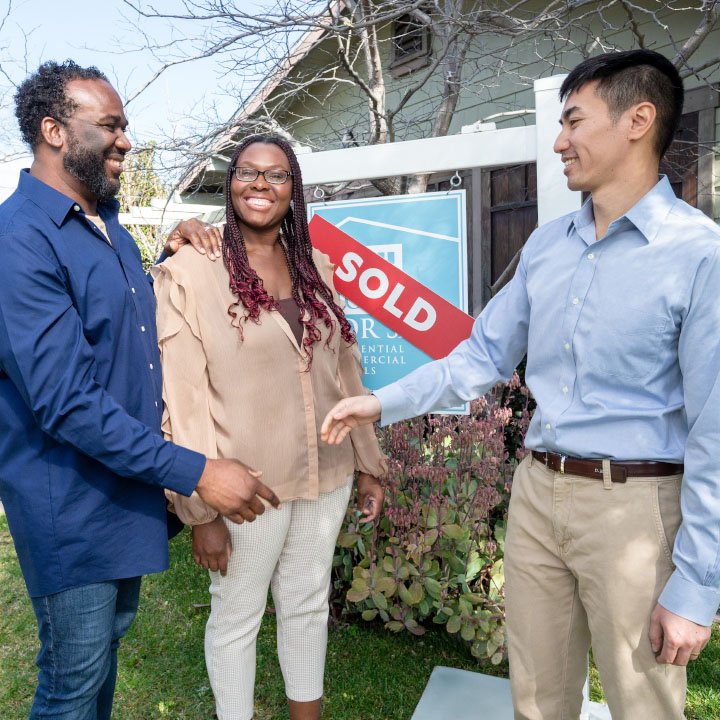 A husband and wife shake hands with a realtor in front of a house they have just purchased.