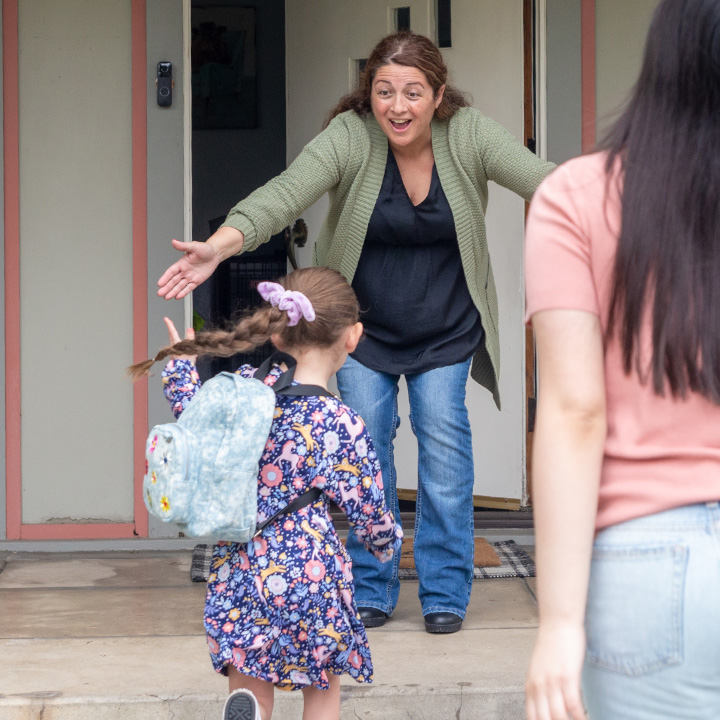 A girl runs to hug a woman standing on a front porch after a day at school.