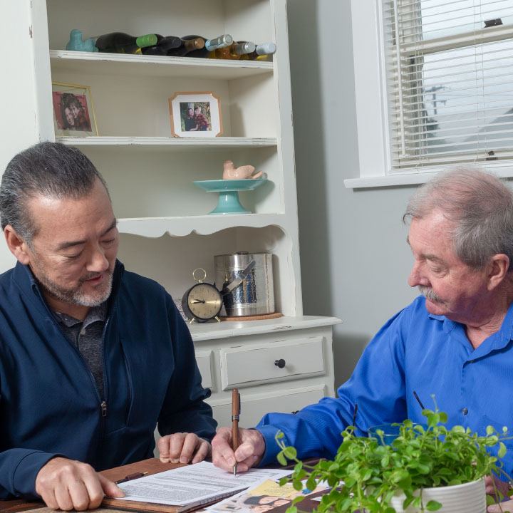 A man with his life insurance agent signs his new TruStage Life Insurance policy at the kitchen table.