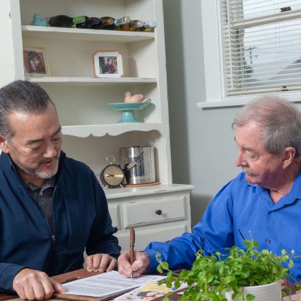 A man with his life insurance agent signs his new TruStage Life Insurance policy at the kitchen table.