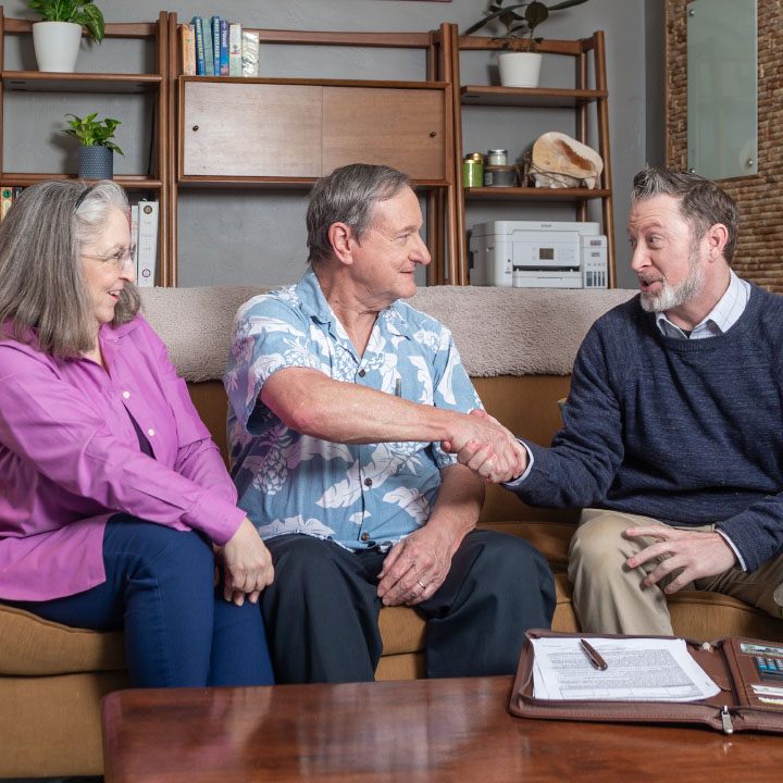 A man shakes hands with his insurance agent while sitting on a couch with his wife.