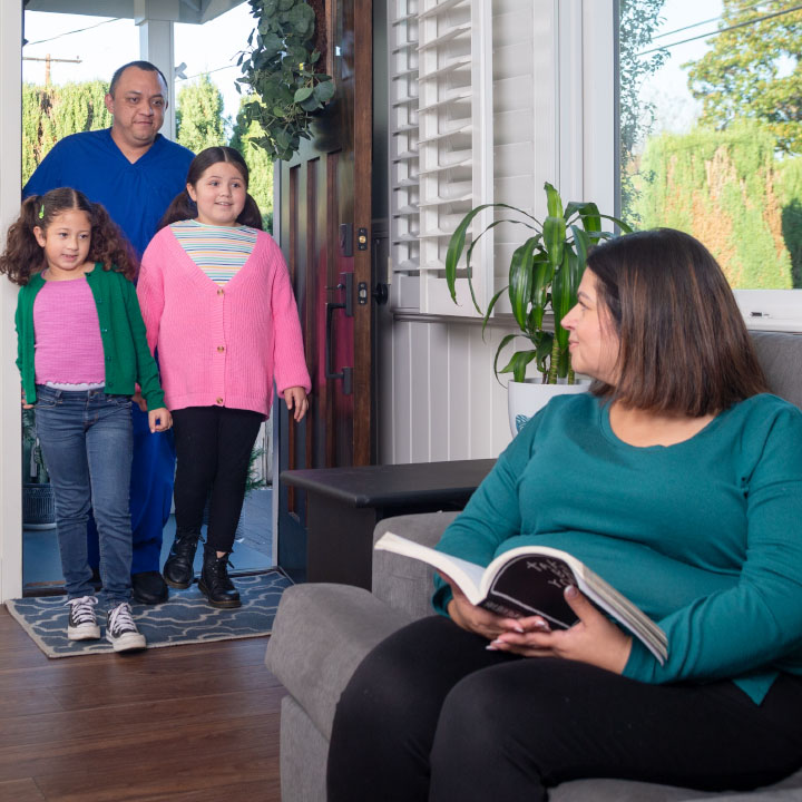 A father walks with his daughters through a front door as his wife reads about living insurance while sitting on a couch.