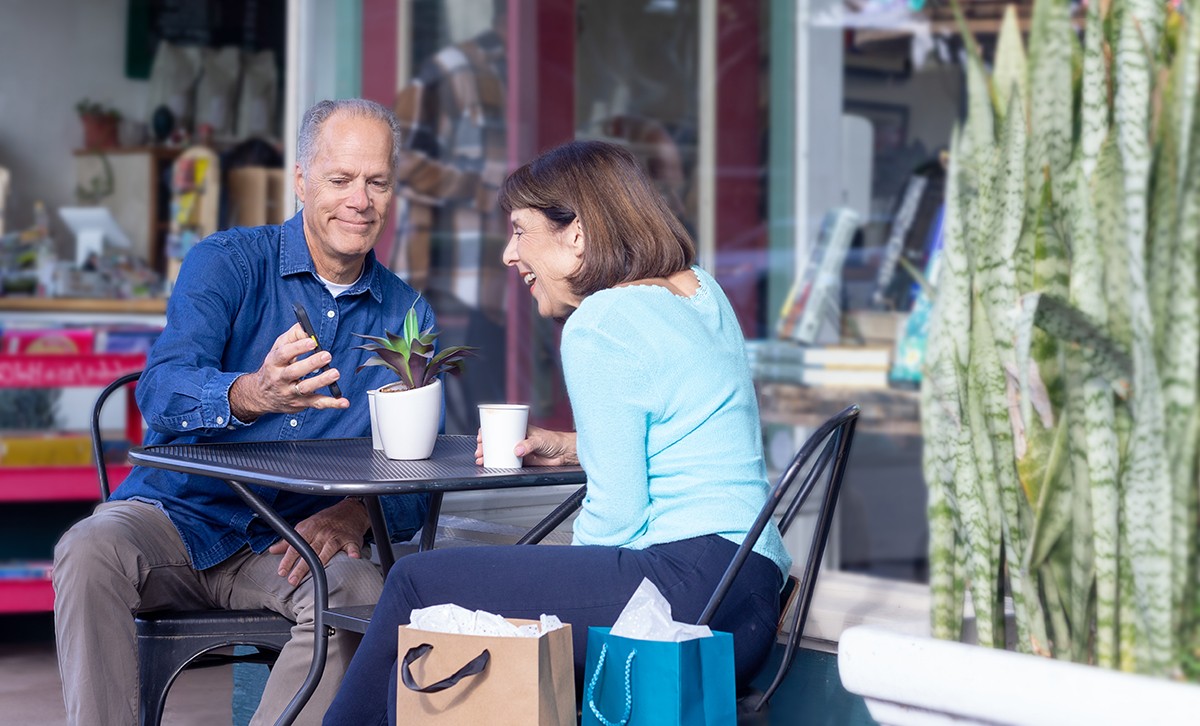 A husband sits with his wife at a table outside a coffee shop showing her a new account he opened using his phone.