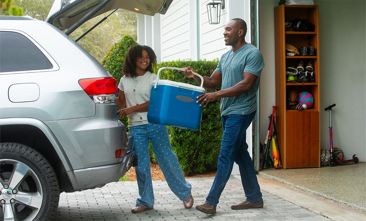 A father and his daughter carry a blue cooler from a garage to the open trunk of an SUV in a driveway.