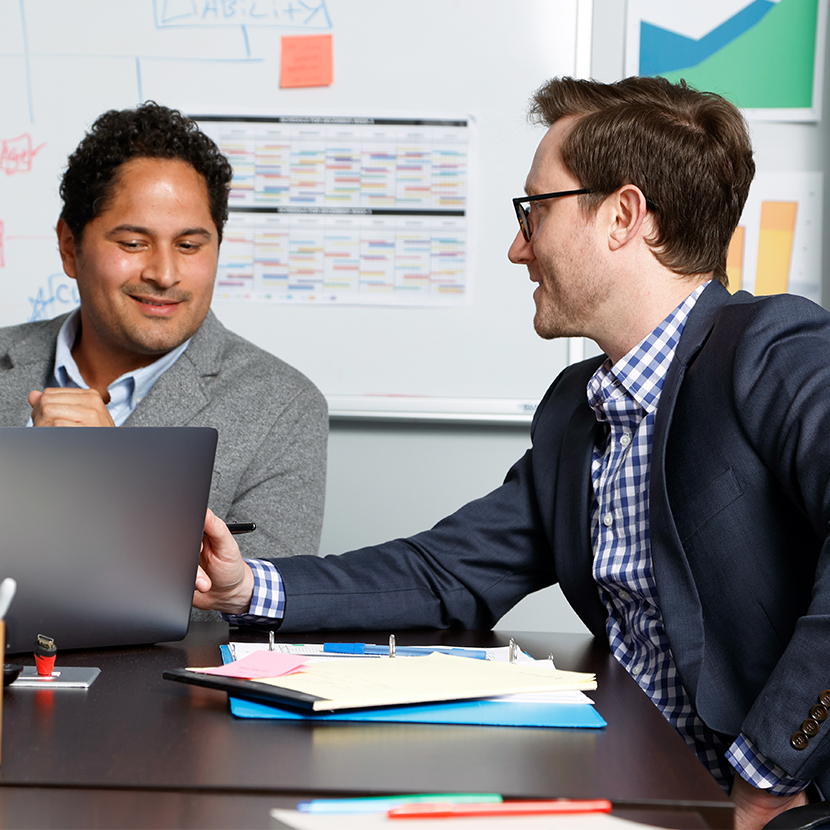 Two men sitting at a desk, one pointing to a computer and the other looking at what he is point to