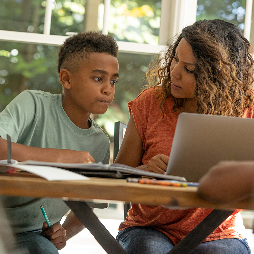 A woman at the table on a laptop sitting next to her son who is writing on a notebook