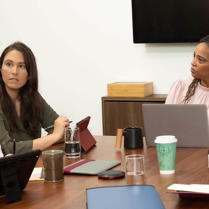 Two women sitting at a desk in an office setting, looking at something offscreen.