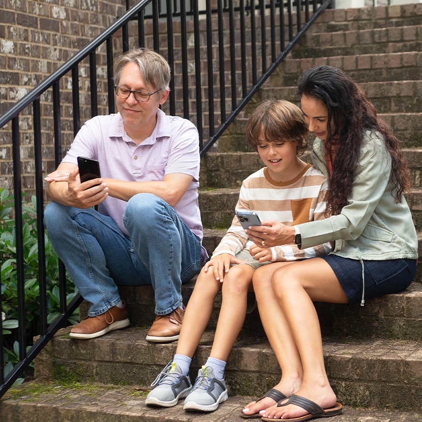 Family sitting on stairs outside with a man looking at a cellphone, and young boy and woman looking at another cellphone.