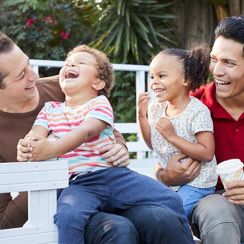 A male couple and their two young children sit in a park feeling thankful for their TruStage life insurance.