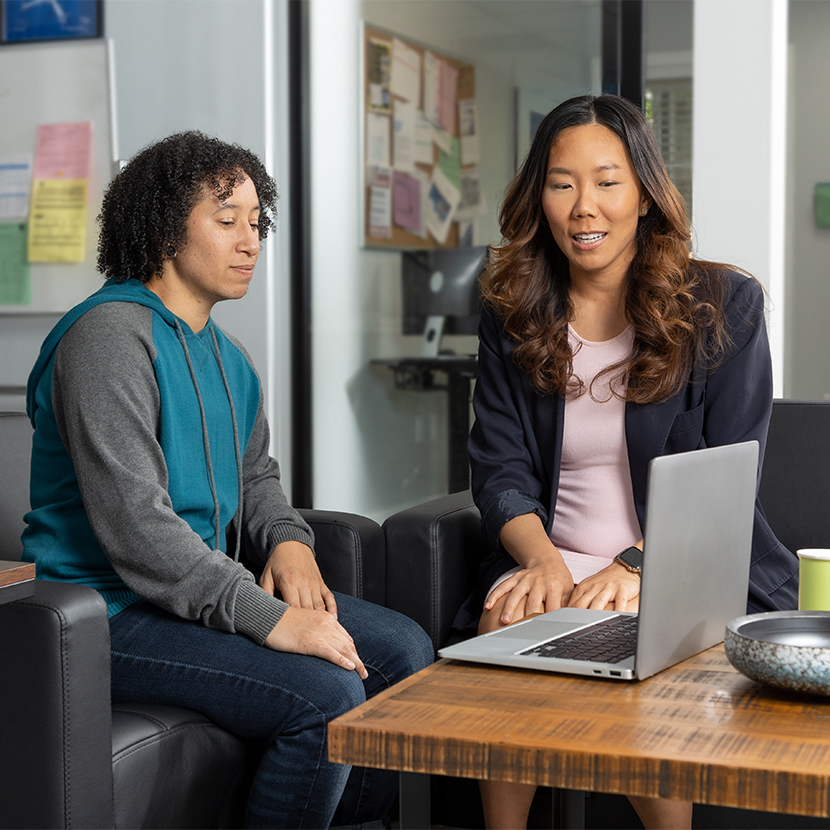 Two women sit in chairs looking at a laptop on the table together