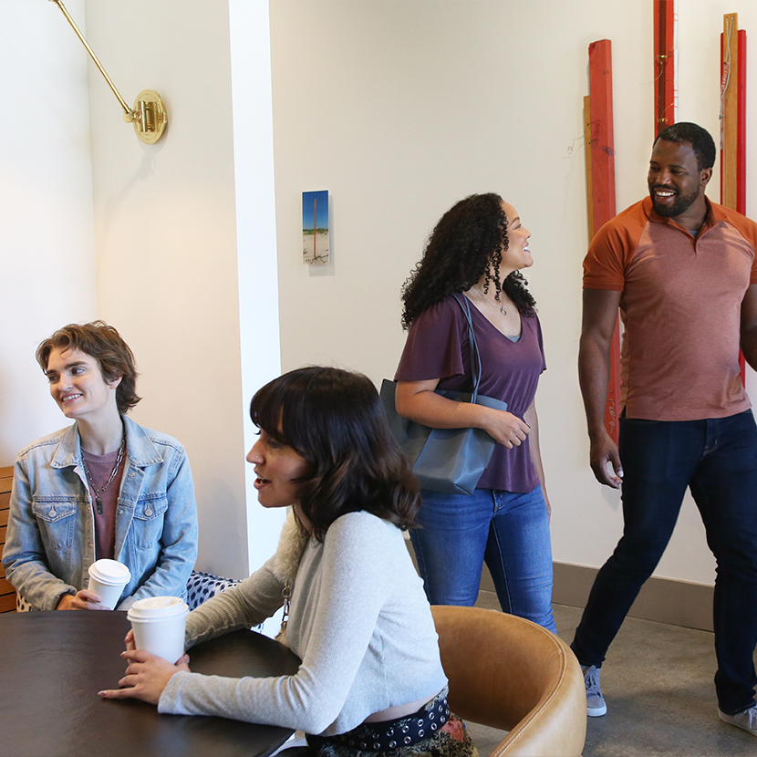 Group of people, two people sitting down at a table with disposable coffee cups in their hands and talking, two people walking and talking