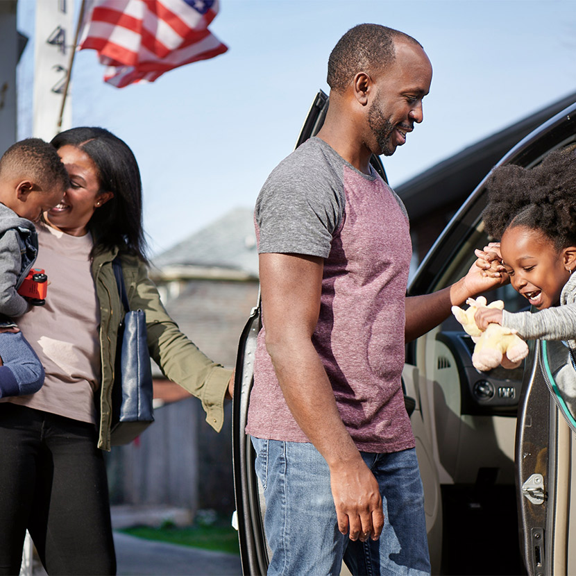 A mother holds her son near a house while the father helps his daughter out of a van