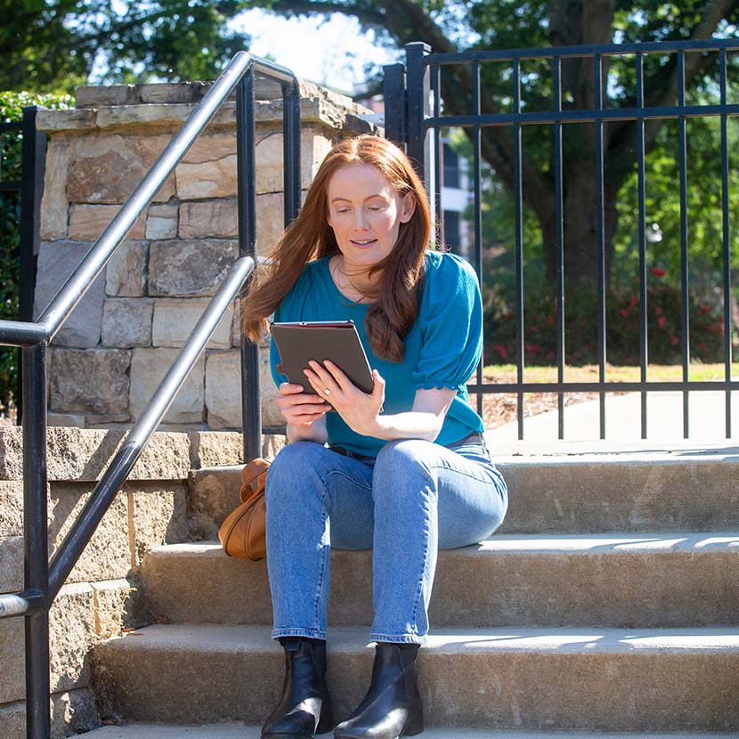 A women seated on some outdoor steps is reading about the IRS 2025 cost-of-living (COLA) updates on her tablet.