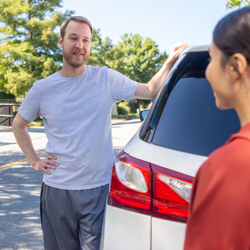 Man holding on the top of a parked car, while talking to a woman on the side of a street