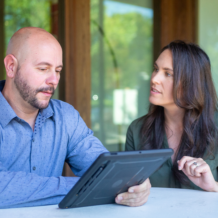 Man and woman sitting at a table, talking, while the man is looking at a tablet