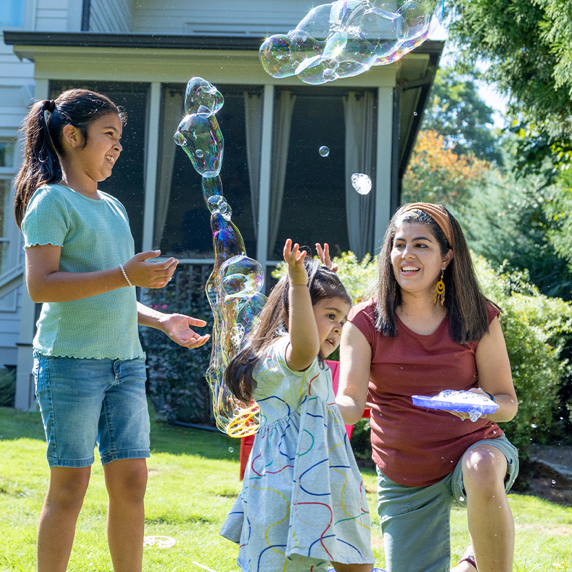 Two young girls and a woman playing with bubbles in front of a house