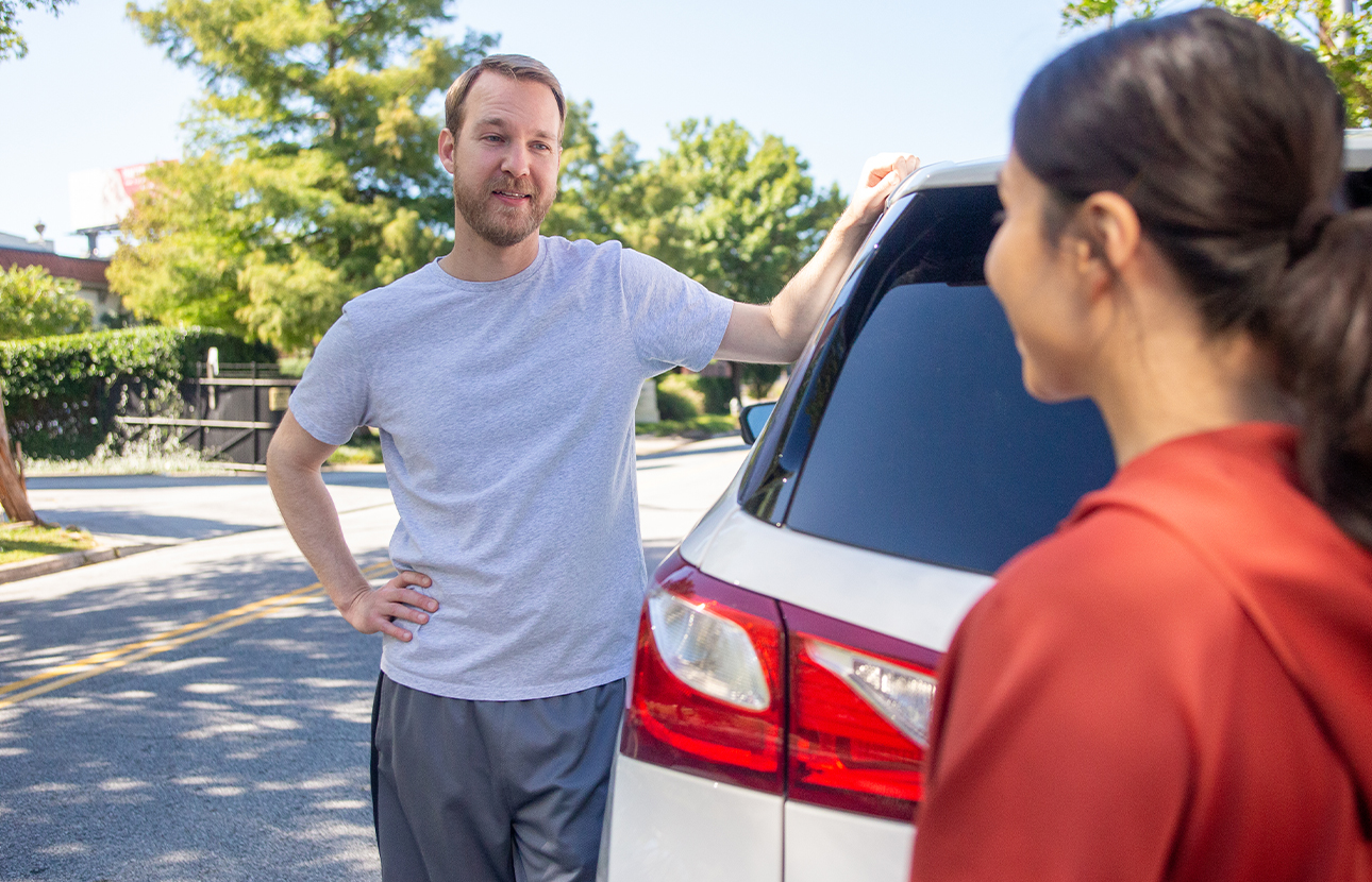 Man holding on the top of a parked car, while talking to a woman on the side of a street