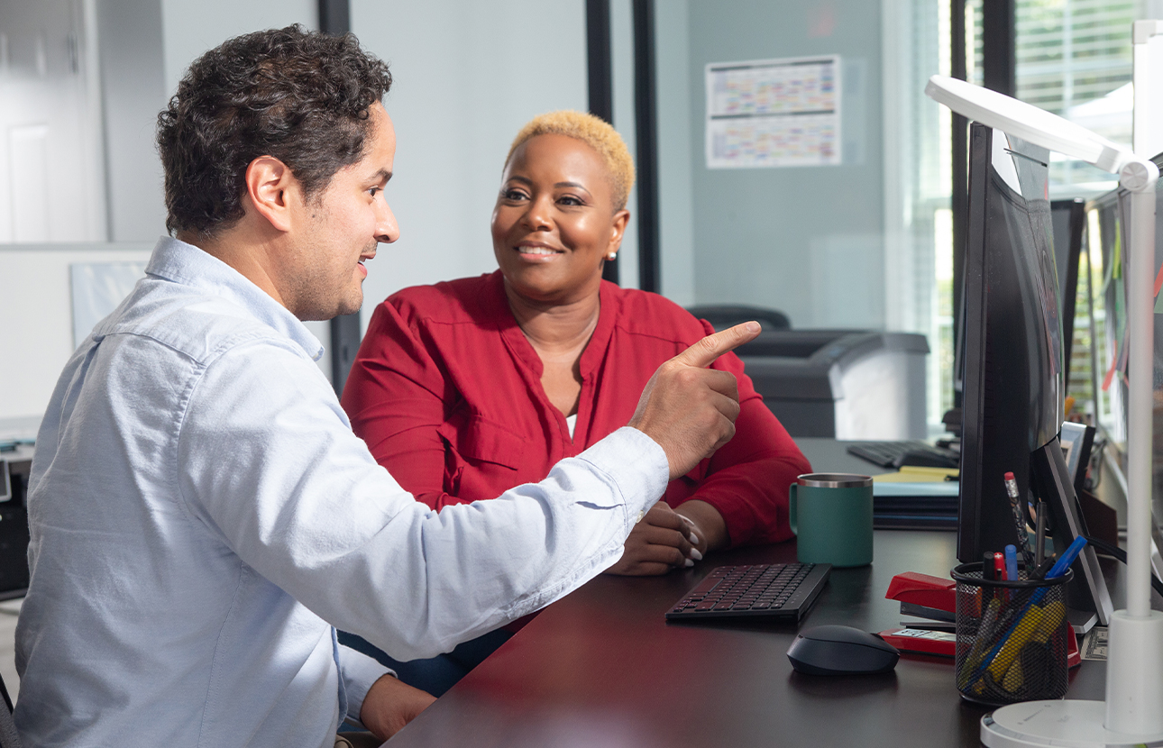 Man and woman talking while seated at a computer desk, man is pointing at the computer screen