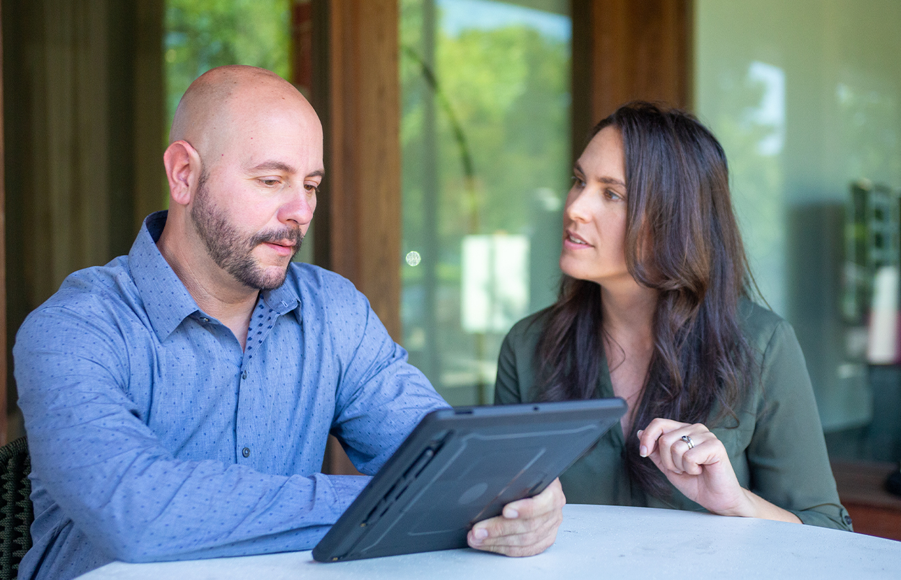 Man and woman sitting at a table, talking, while the man is looking at a tablet