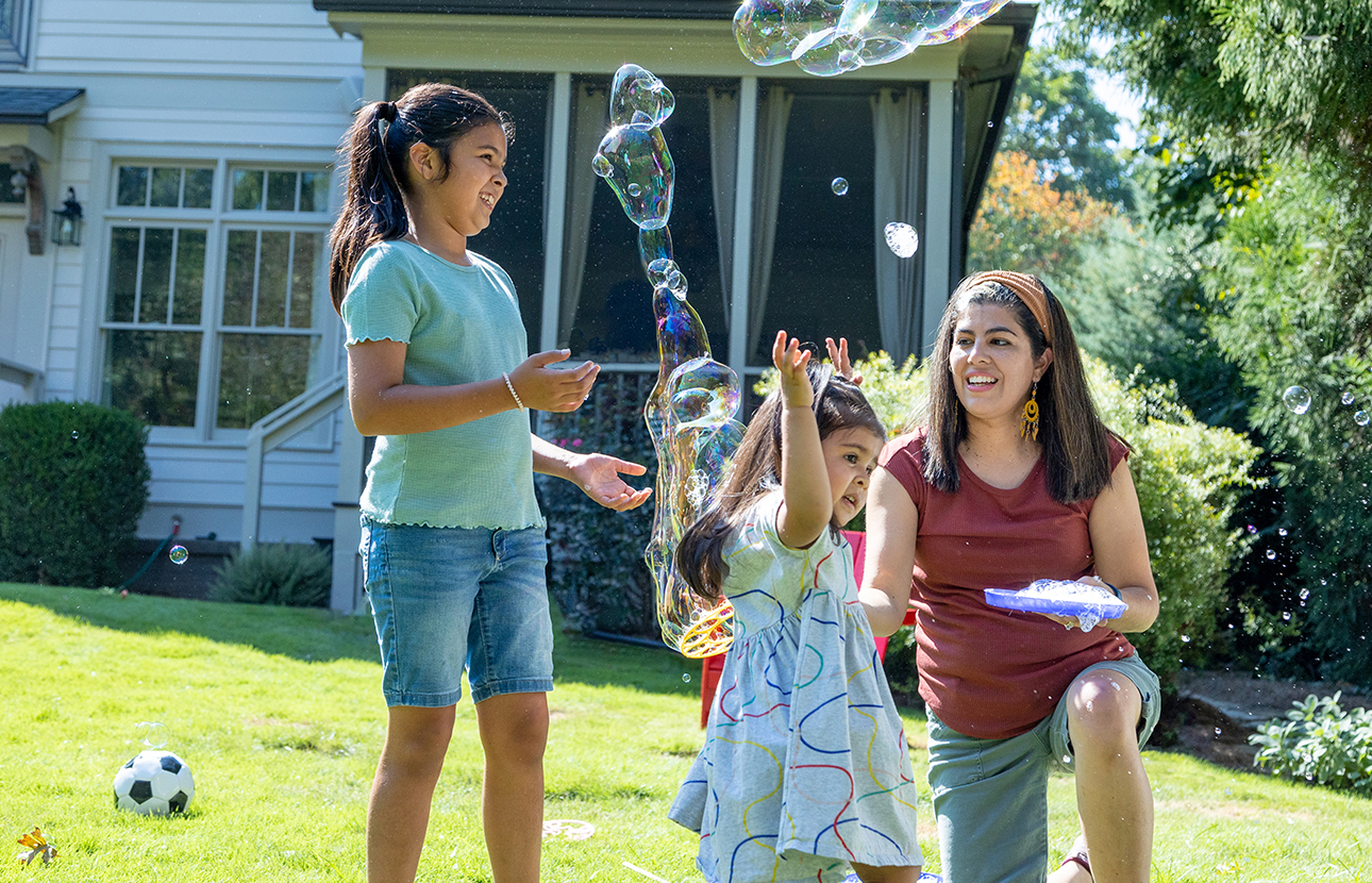 Two young girls and a woman playing with bubbles in front of a house