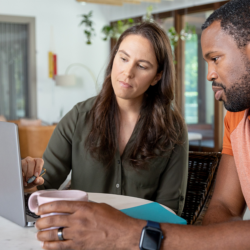 Woman and man sitting at a table looking at a laptop, woman holding a pen and man holding a coffee mug