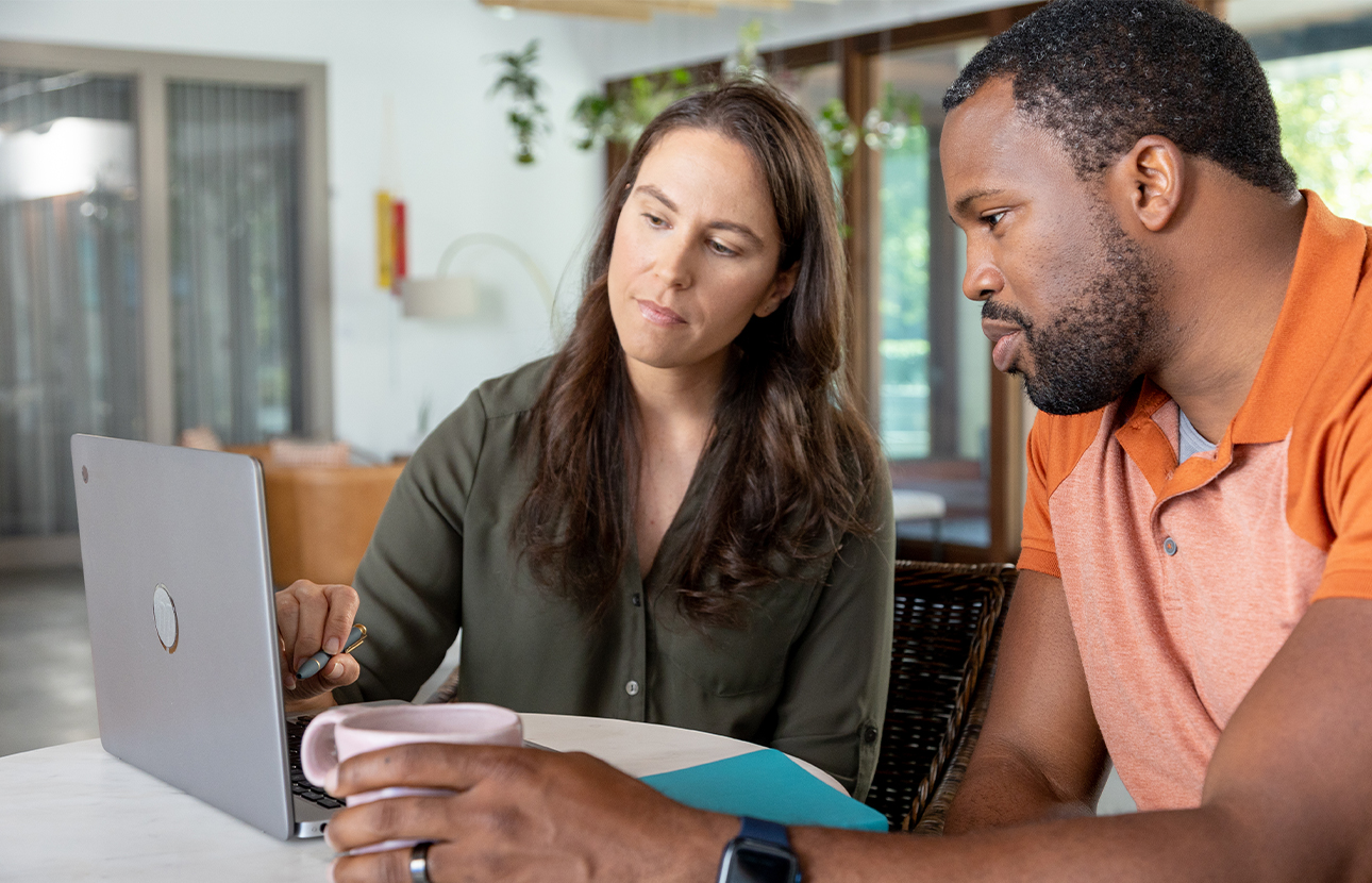 Woman and man sitting at a table looking at a laptop, woman holding a pen and man holding a coffee mug