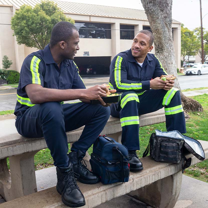 Two workers in dark uniforms with reflective stripes sit on a bench eating lunch outdoors.