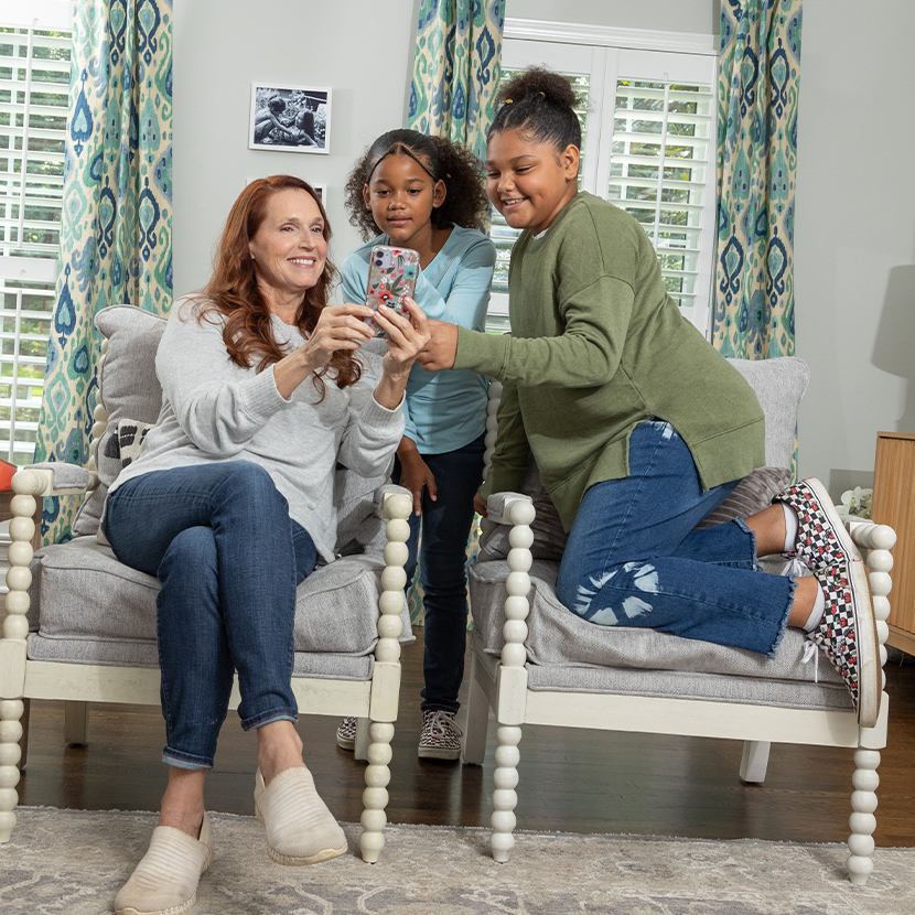 A woman and two young girls in a house, the woman and one girl sitting in chairs, while the other girl is standing, looking at a cell phone