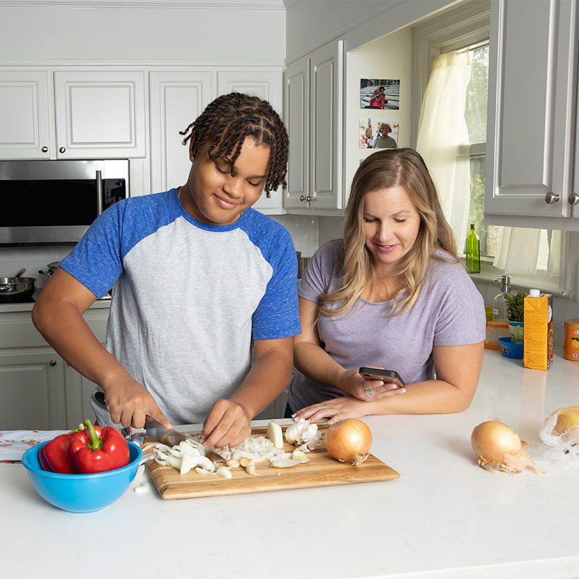 Woman and an older boy in a kitchen, the woman is on her cellphone and the boy is chopping onions