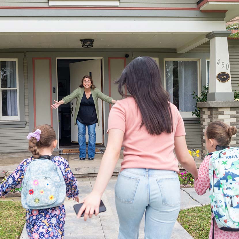 An adult with two kids wearing backpacks walks toward a house where someone stands with open arms.