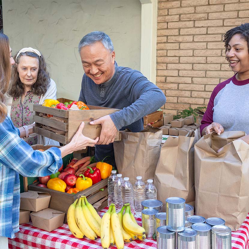 People gather around a table with fresh produce, canned goods, and paper bags at an outdoor food stand.