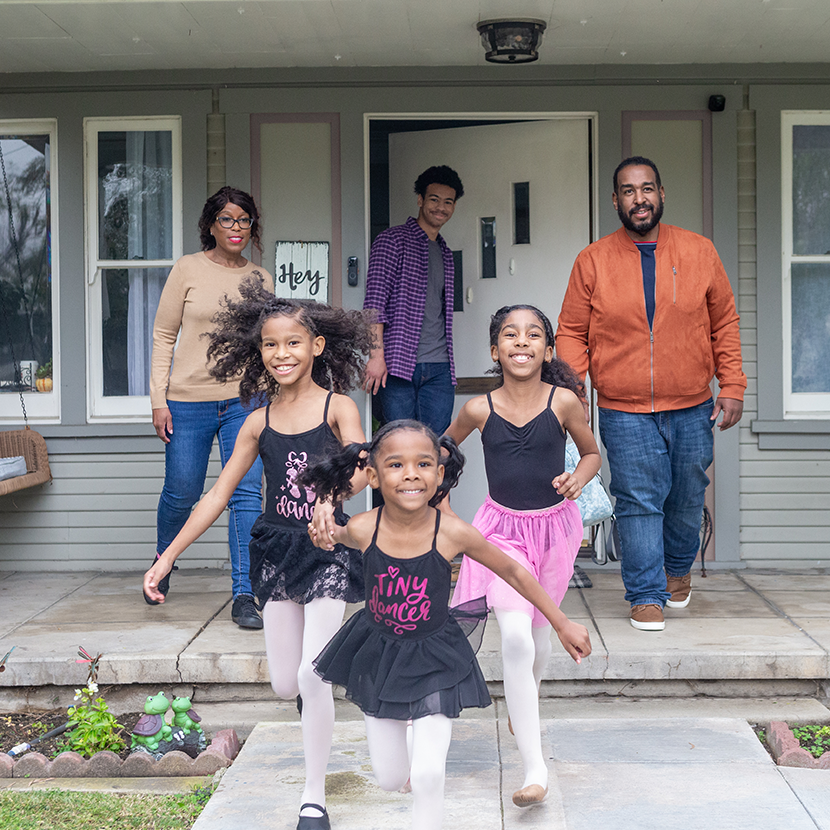 Three kids in ballet outfits run on a porch while three adults stand in front of a gray house with flowers.
