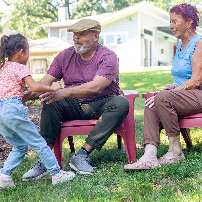 Man and woman sitting outside in chairs, while a young girl is walking towards the man with hands out and about to be picked up