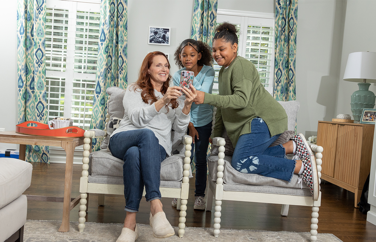 A woman and two young girls in a house, the woman and one girl sitting in chairs, while the other girl is standing, looking at a cell phone