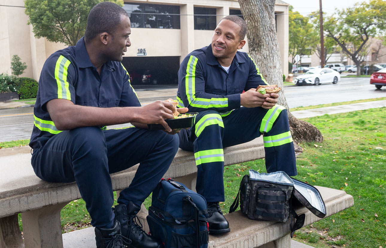 Two men in black jumpsuits with reflective strips on the arms and legs are sitting on a picnic table talking to each other while they eat food out of lunch bags.