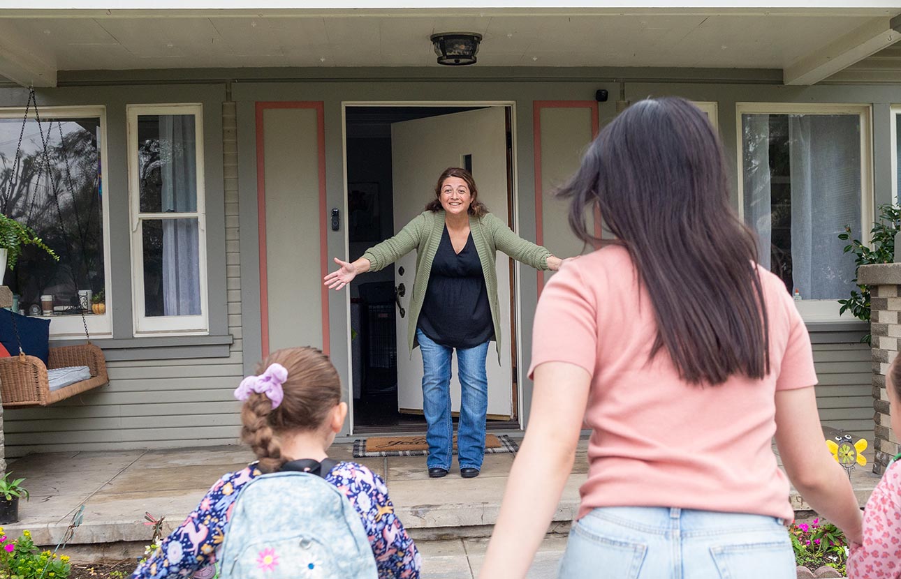 Adult with kid wearing backpack walks toward a house where someone stands with open arms.