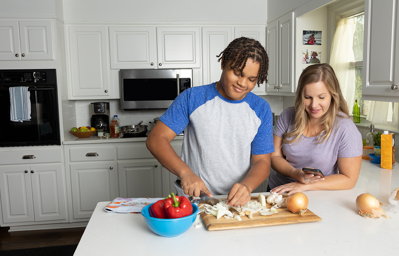 Woman and an older boy in a kitchen, the woman is on her cellphone and the boy is chopping onions
