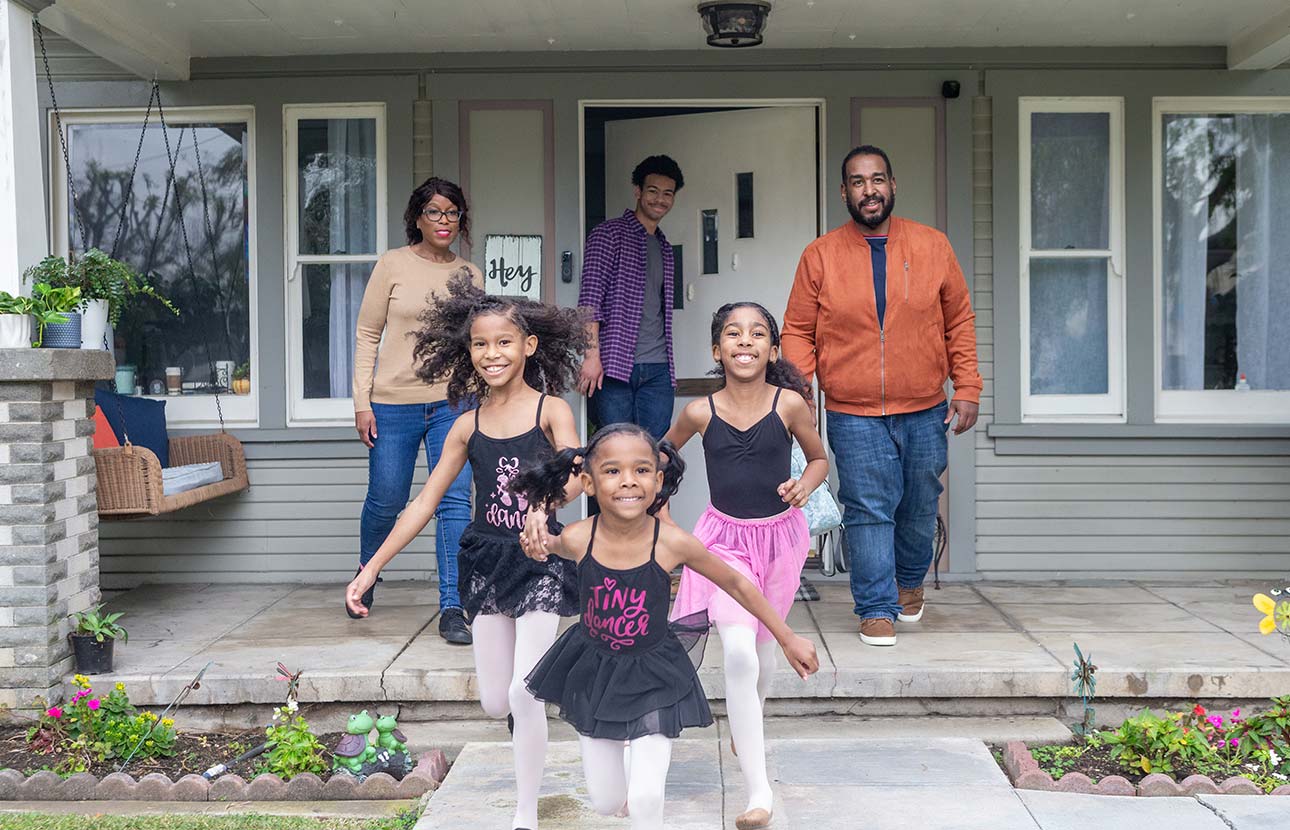 Three kids in ballet outfits run on a porch while three adults stand in front of a gray house with flowers.